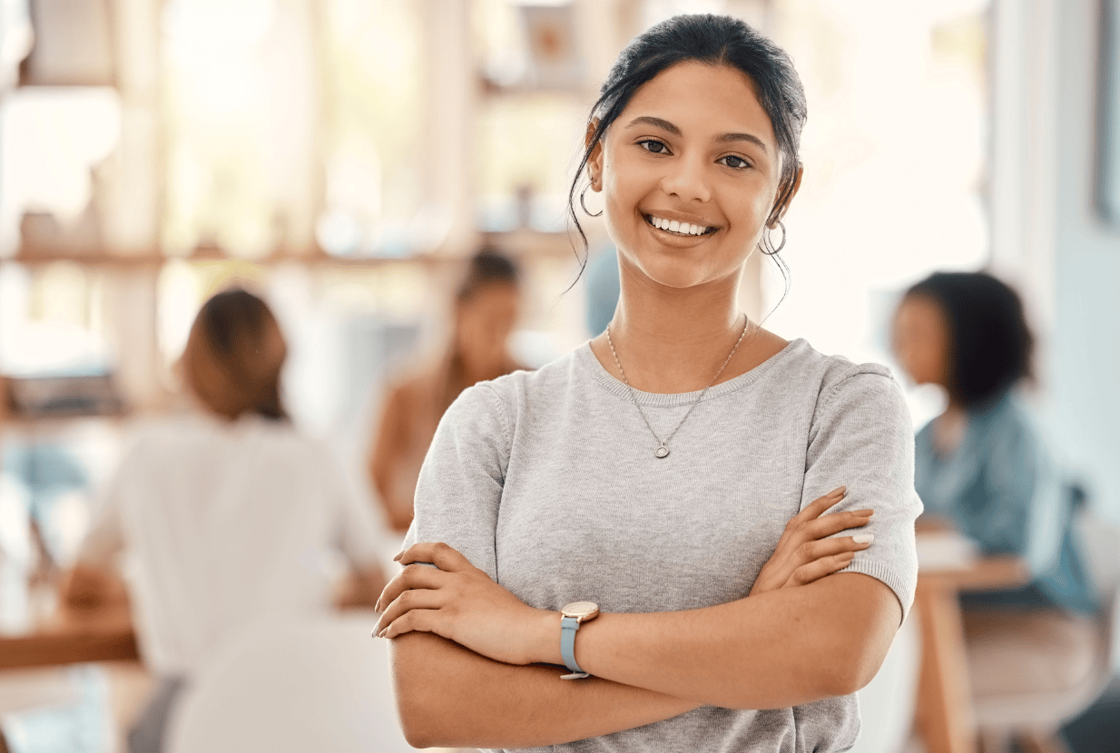 A young woman with dark hair smiles confidently with arms crossed, standing in a bright office. Other people sit and talk in the blurred background.