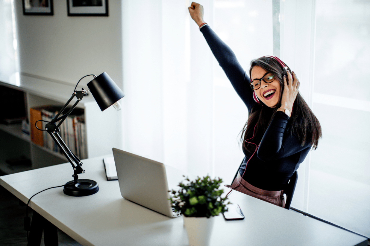 A woman wearing headphones sits at a desk with a laptop and lamp, smiling and raising one arm in excitement, in a bright, modern room.