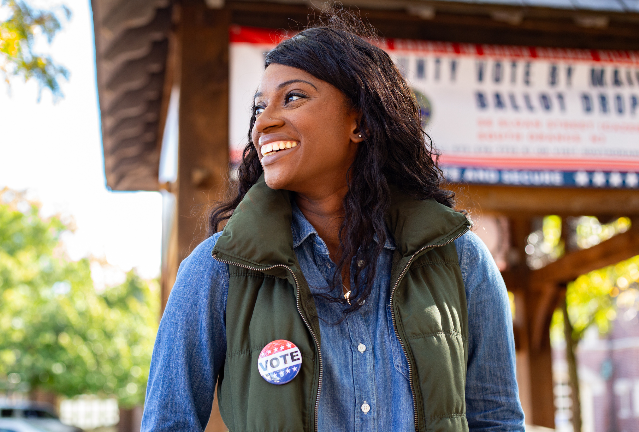 Smiling woman wearing a VOTE button stands outdoors near a ballot drop box location, with a banner about mail-in voting in the background.