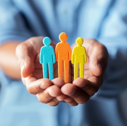 A person holds three colorful cutouts of human figures—blue, orange, and yellow—in their cupped hands, symbolizing diversity, support, and unity.