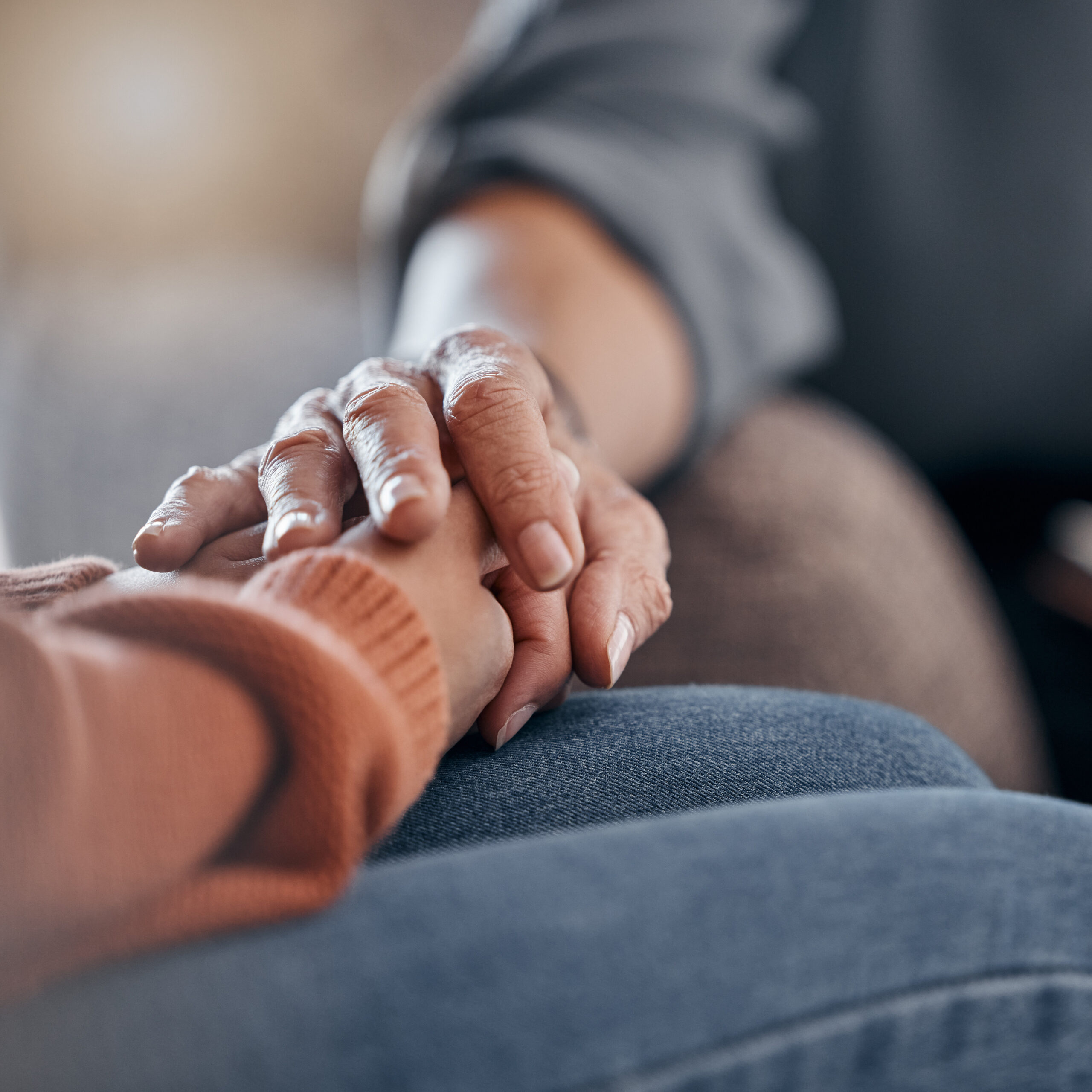 A close-up of two people sitting side by side, with one person gently holding the others hand for comfort and support. Both wear casual clothing, and the scene conveys empathy and care.