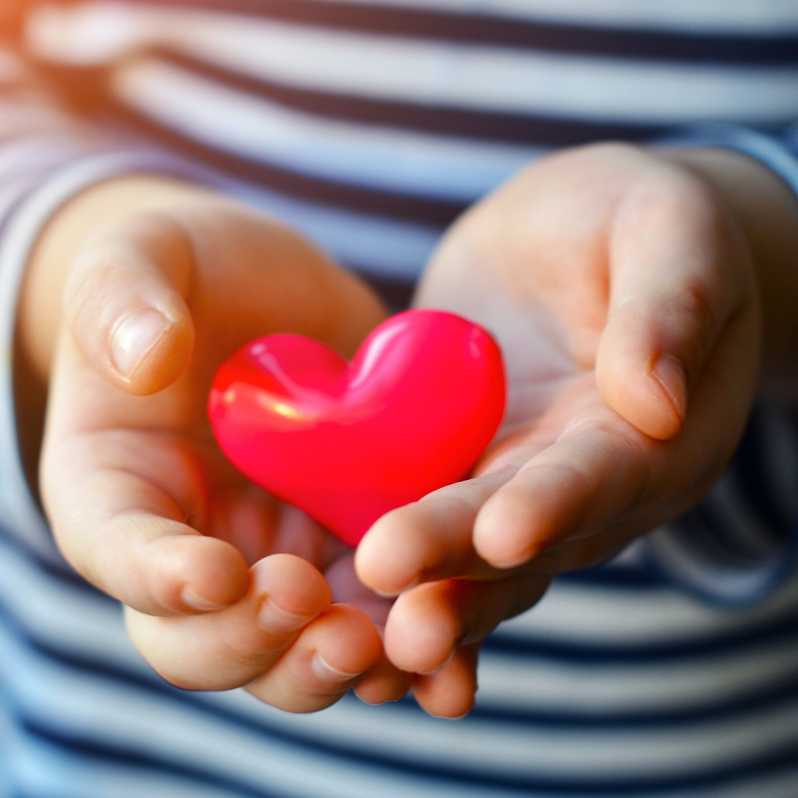 A person in a striped shirt holds a shiny red heart-shaped object gently in their open hands.