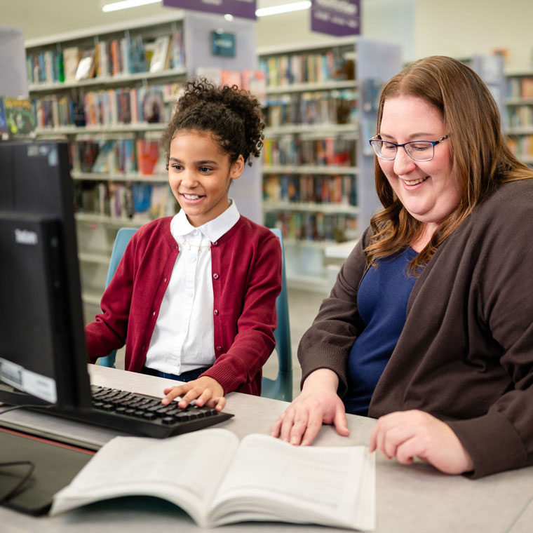 A smiling girl and woman sit together at a computer in a library, with bookshelves in the background and an open book on the desk.
