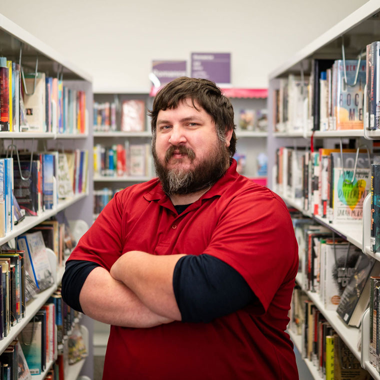 A person with a beard and mustache, wearing a red polo shirt, stands with arms crossed between library bookshelves filled with books.