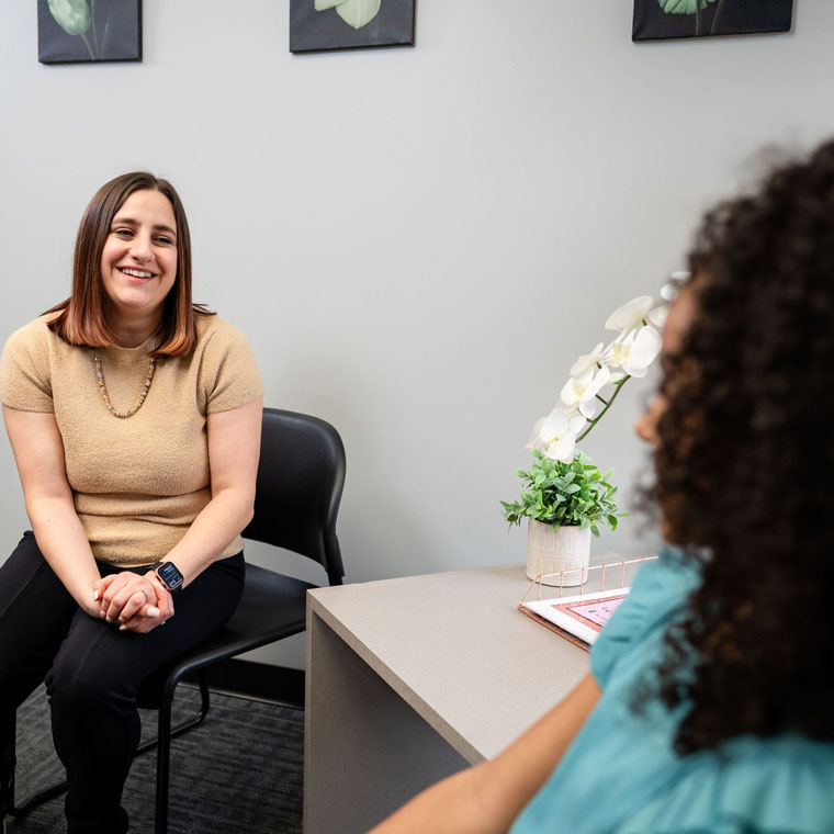 A woman sits and smiles while talking to another person across a desk in an office with light walls and framed pictures.
