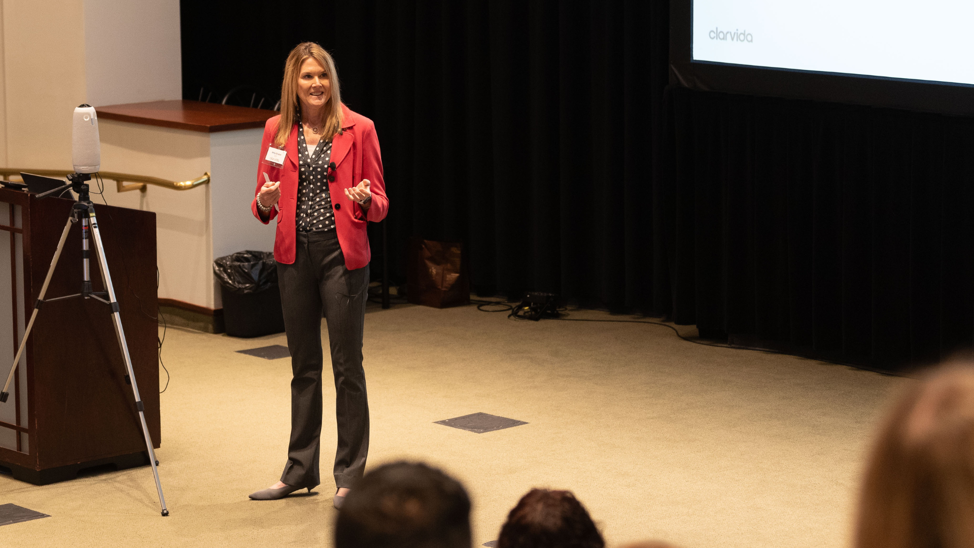 A woman in a red blazer gives a presentation in a conference room, standing near a podium and a tripod. Audience members are visible in the foreground.