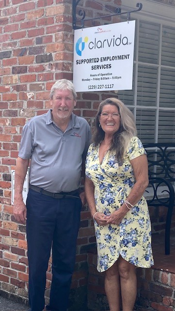 Two adults stand smiling in front of a brick building with a sign reading clarvida Supported Employment Services. The man wears a gray shirt and dark pants; the woman wears a yellow floral dress.
