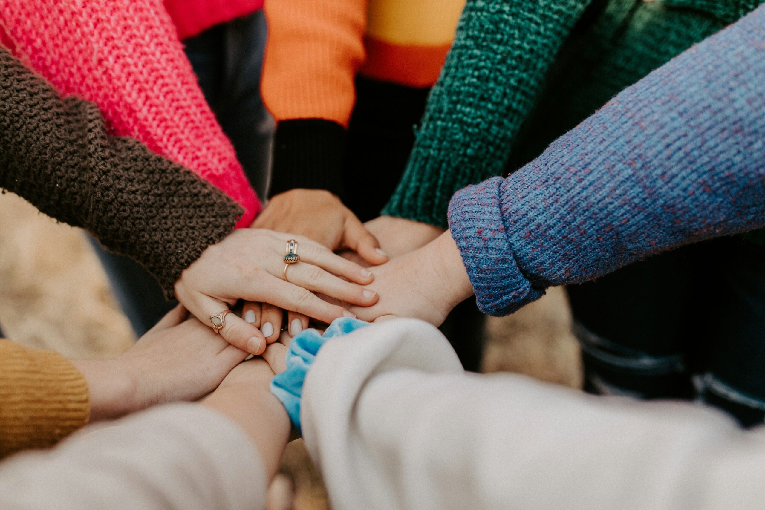 A group of people wearing colorful sweaters place their hands together in a circle, symbolizing unity and teamwork.