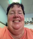 A person with short dark hair smiles at the camera while wearing a peach shirt. The background shows a light blue wall and ceiling, with some signs or labels slightly visible behind them.