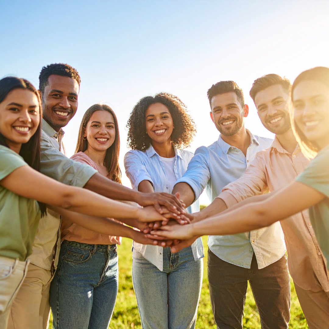 A group of seven people outdoors stands in a circle, smiling and stacking their hands together in the center, showing teamwork and unity. The sun is shining brightly in the background.