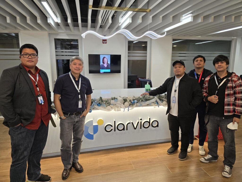 Five people stand in front of a reception desk with the Clarvida logo, wearing ID lanyards. The modern office has a decorated counter and a TV screen on the wall behind them displaying a womans face.