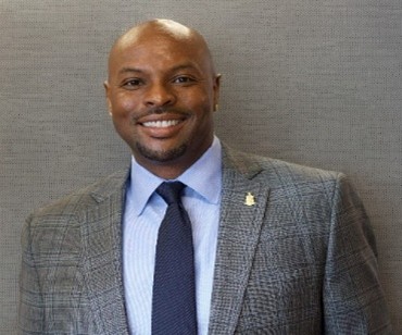 A man in a plaid gray suit, light blue shirt, and navy tie stands smiling in front of a plain gray background.
