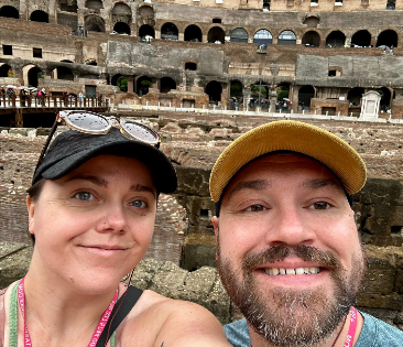 Two people smile for a selfie in front of the Colosseum in Rome. Both wear caps and pink lanyards, with ancient stone walls and arches visible in the background.