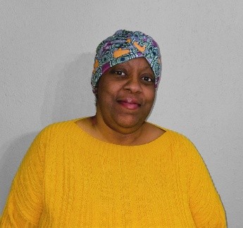 A woman wearing a patterned headscarf and a bright yellow sweater stands in front of a plain gray wall, smiling slightly at the camera.