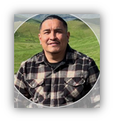 A person wearing a black and white plaid shirt is smiling and posing outdoors with green hills in the background.