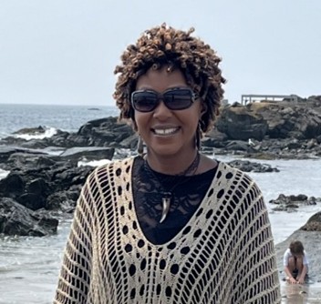 A woman wearing sunglasses and a light knit sweater smiles while standing on a rocky beach, with the ocean and rocks visible in the background.