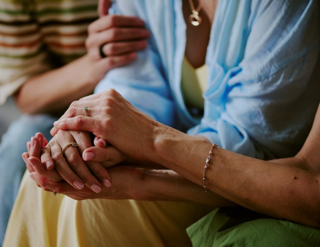 Two people comfort each other, with one person gently holding and supporting the others hands. Both appear to be sitting close, wearing casual clothing and jewelry, creating a caring and supportive atmosphere.