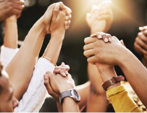 A group of diverse hands are raised and clasped together in unity outdoors, with sunlight shining in the background.