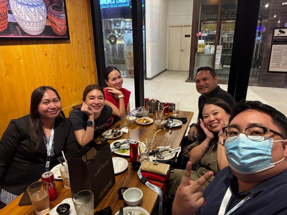 Six people sit around a restaurant table with empty plates and drinks, smiling at the camera. One person in the foreground wears a face mask and makes a peace sign.