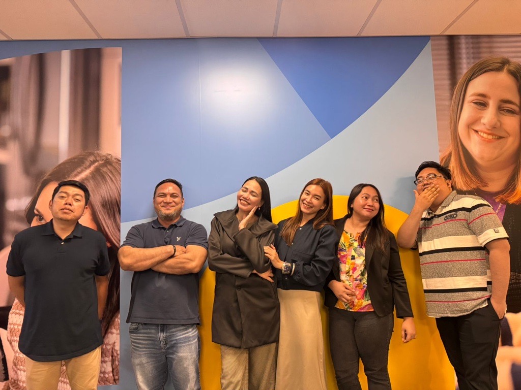 Six people stand in a row in front of a colorful wall, posing and smiling playfully. Two large portraits are partially visible in the background on the left and right sides.