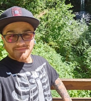 A person wearing glasses, a cap, and a skeleton-print shirt smiles on a sunny day, standing by a wooden railing with lush green trees and a small waterfall in the background.