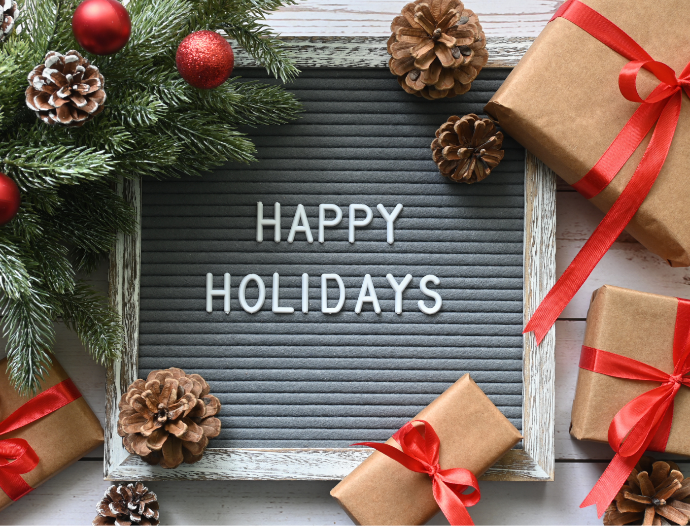 A letter board with HAPPY HOLIDAYS is surrounded by pine branches, red ornaments, pinecones, and brown gift boxes tied with red ribbons on a white wooden surface.