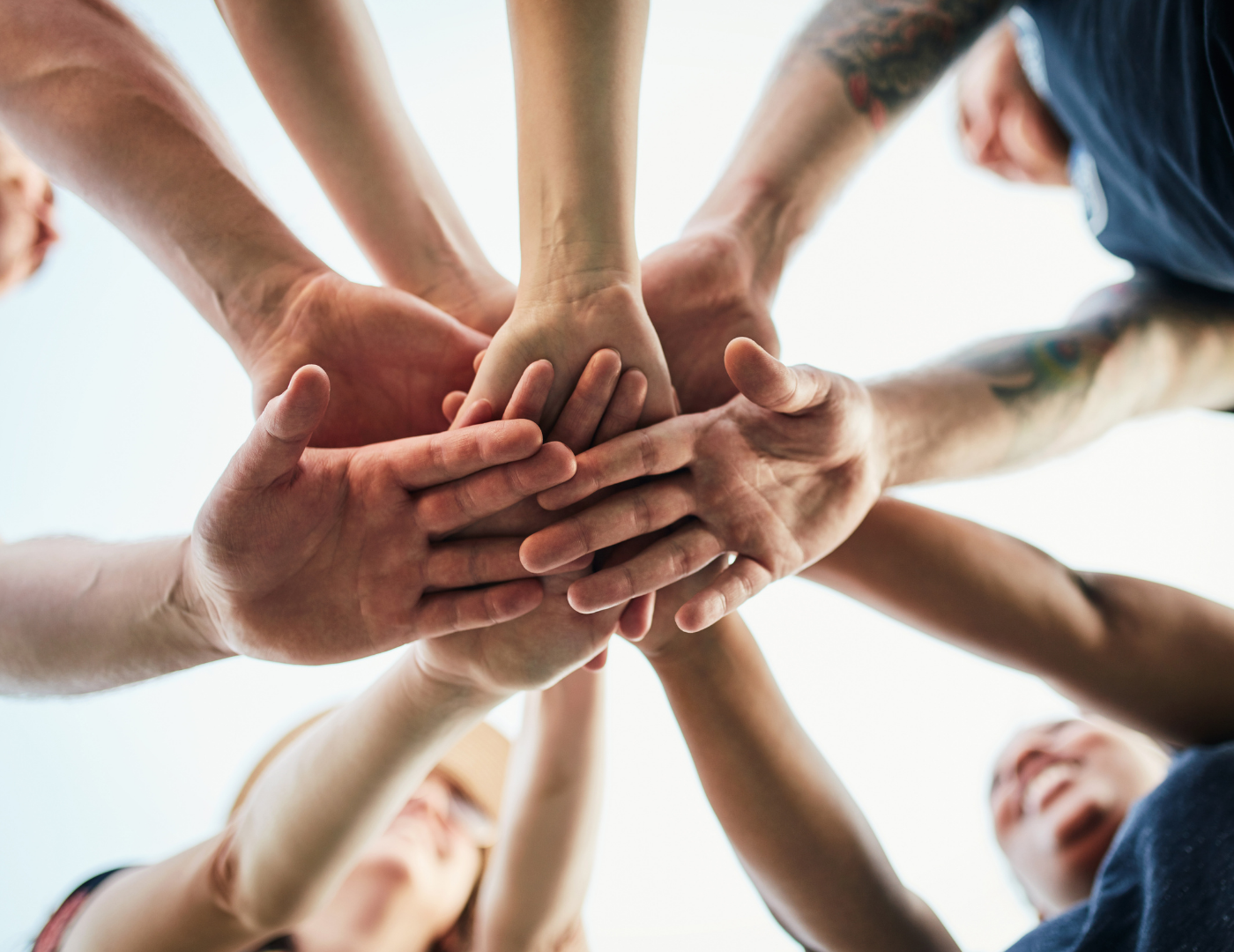 A group of people stand in a circle, reaching into the center to stack their hands together, symbolizing unity and teamwork, with smiling faces visible in the background.