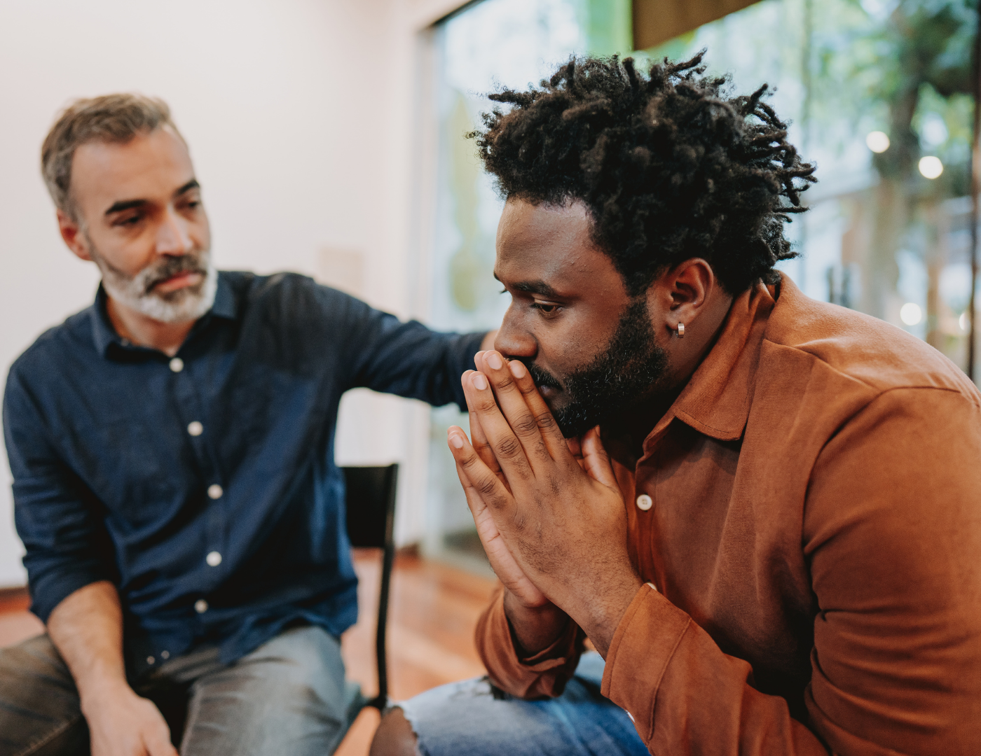 Two men sitting indoors; one looks concerned with hands clasped by his face, while the other gently touches his shoulder, offering comfort and support.