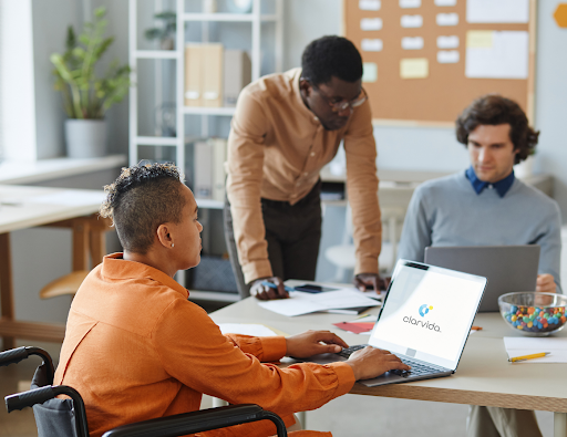 Three people in an office work together at a table, demonstrating disability inclusion in the workplace. One person in a wheelchair uses a laptop displaying the Clarvida logo, while the others discuss documents.