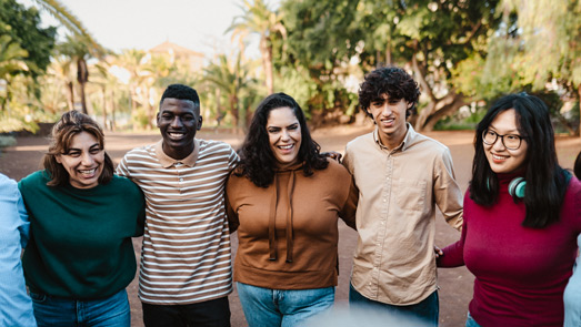 A group of five people stand outdoors with arms around each other, smiling and looking happy. Their sense of connection and joy highlights the importance of behavioral health, while trees and greenery in the background suggest a park or garden setting.