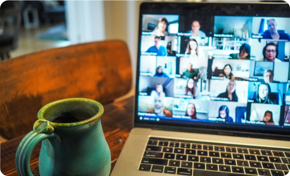 A green mug sits on a wooden table next to a laptop displaying a video conference with many people in small squares on the screen.